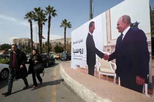 لافتة كبيرة تظهر بشار الأسد وفلاديمير بوتين أثناء مصافحة في سياق دعم روسيا لنظام الأسد في سوريا، مع أشجار النخيل والسيارات في الخلفية.