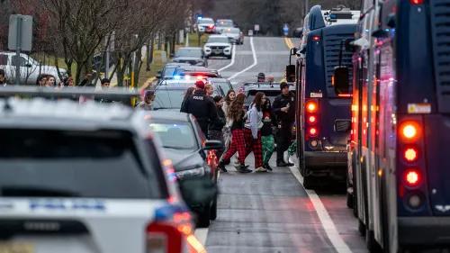 طلاب يخرجون من مدرسة بعد حادث إطلاق نار، مع وجود ضباط شرطة ومركبات طوارئ في الخلفية، مشهد يعكس حالة من الفوضى والقلق.