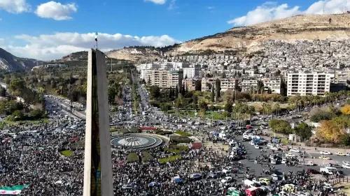 حشود كبيرة من المتظاهرين في ساحة بمدينة سورية، مع العلم الوطني يرفرف، وسط تواجد كثيف للمركبات والمباني المحيطة.