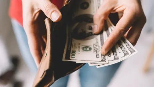يد تُخرج نقودًا من محفظة جلدية، مع التركيز على الأوراق النقدية من فئة المئة دولار، في سياق الحديث عن دخل الأسر الأمريكية.