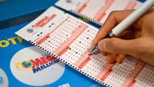 شخص يملأ بطاقة يانصيب Mega Millions، مع ظهور شعار اليانصيب وأرقام السحب على الطاولة، استعدادًا للسحب القادم.