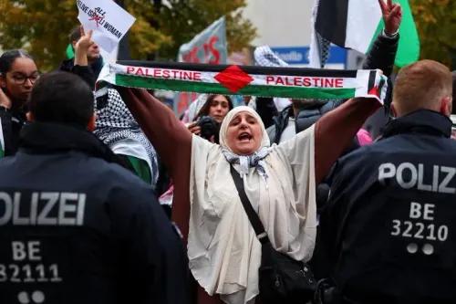 امرأة ترتدي حجابًا وتحمل وشاحًا مكتوبًا عليه \"فلسطين\" وسط حشد من المتظاهرين، بينما يتواجد رجال الشرطة في الخلفية.