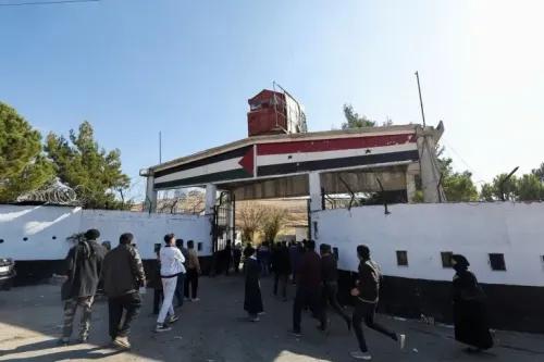 مدخل سجن سيء السمعة في سوريا، حيث يتجمع عدد من الأشخاص أمام البوابة، ويظهر العلم السوري على المبنى، مما يرمز إلى الانتهاكات الحقوقية.