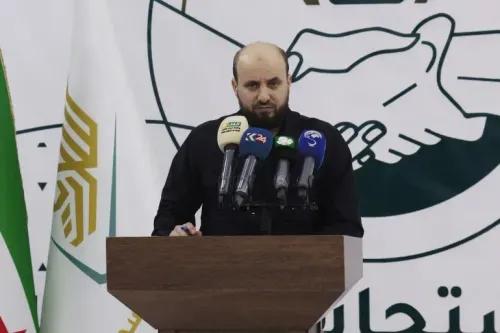 محمد البشير يتحدث في مؤتمر صحفي بعد تعيينه رئيسًا لحكومة تصريف الأعمال في سوريا، مع خلفية شعار الحكومة.