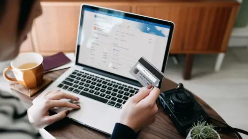 امرأة تحمل بطاقة ائتمان وتستخدم الكمبيوتر المحمول لشراء تذكرة طيران، مع وجود كوب شاي وكاميرا على الطاولة.