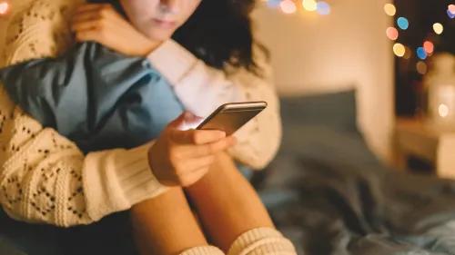 امرأة تجلس على السرير تحتضن وسادة، تستخدم هاتفها المحمول في غرفة مضاءة بأضواء خافتة، تعكس قلقها بشأن الصحة النفسية.