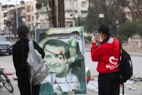 صورة لفتاتين تقفان أمام لوحة مشوهة لبشار الأسد في شوارع دمشق، تعكس أجواء الاحتفالات بعد تقدم المعارضة السورية.