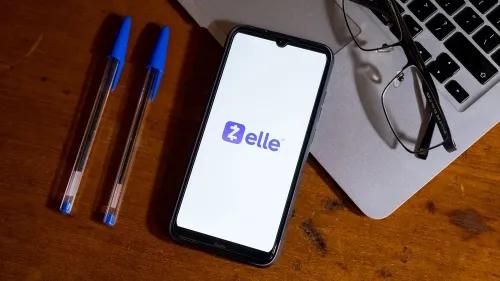 شاشة هاتف تعرض شعار Zelle مع قلمين وزجاجة نظارات على سطح خشبي، تشير إلى مشاكل في خدمة الدفع.